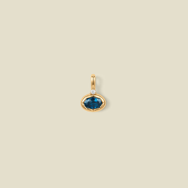 A gold oval pendant charm with a blue-green bezel-set oval gemstone and a clear accent stone on top, set against a solid beige background.