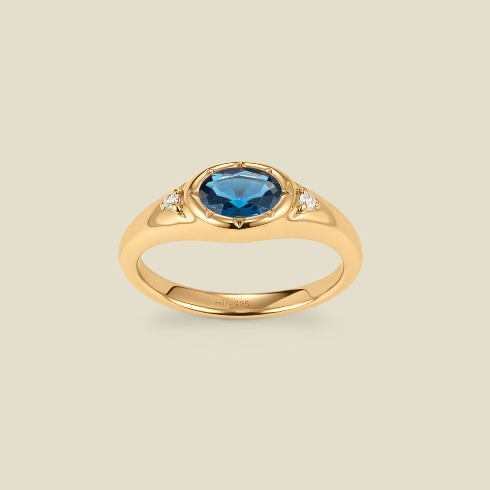 A gold signet ring with a blue-green center oval bezel-set gemstone, and two small clear accent stones on either side, set against a solid beige background.