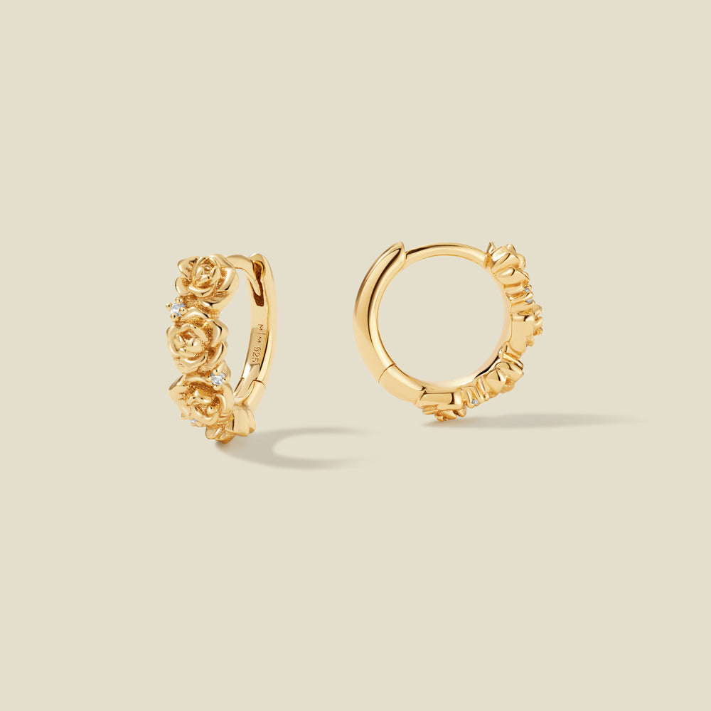 A pair of gold hoop earrings adorned with textured peony flowers stacked along the hoop, with scattered small clear accent stones, set against a solid beige background.
