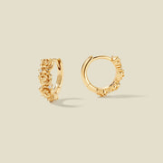 A pair of gold hoop earrings adorned with textured peony flowers stacked along the hoop, with scattered small clear accent stones, set against a solid beige background.