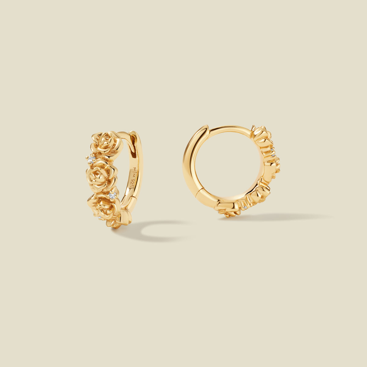 A pair of gold hoop earrings adorned with textured peony flowers stacked along the hoop, with scattered small clear accent stones, set against a solid beige background.
