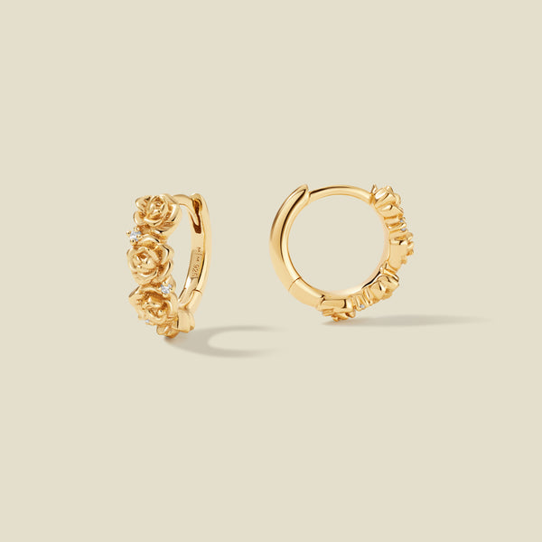 A pair of gold hoop earrings adorned with textured peony flowers stacked along the hoop, with scattered small clear accent stones, set against a solid beige background.