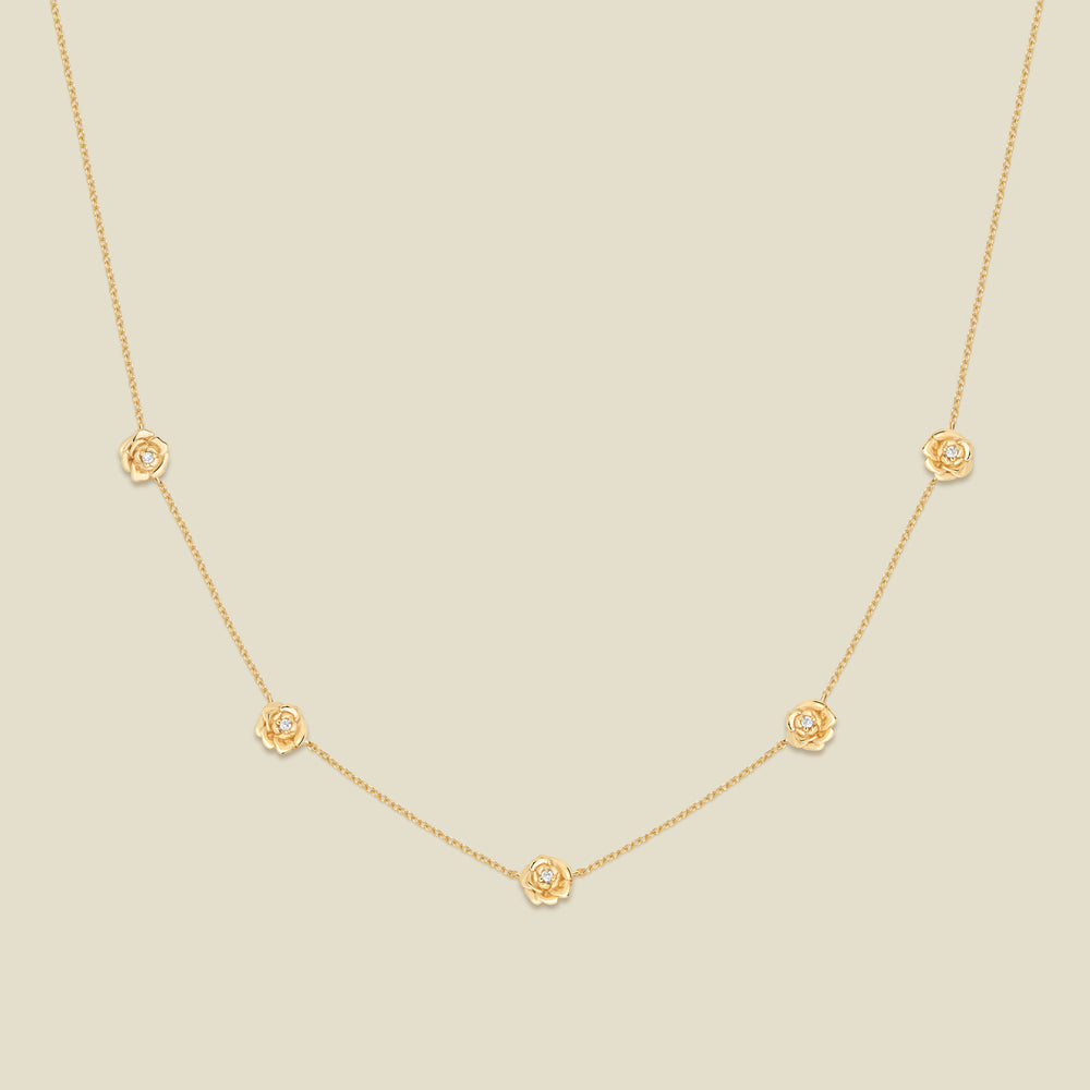 A delicate gold chain necklace featuring five small, evenly spaced textured peony flower motifs set with tiny clear stones, displayed on a solid light beige background.