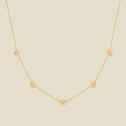 A delicate gold chain necklace featuring five small, evenly spaced textured peony flower motifs set with tiny clear stones, displayed on a solid light beige background.