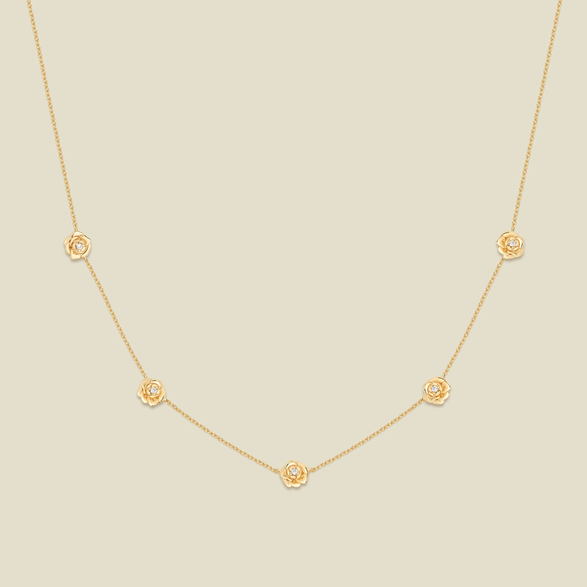 A delicate gold chain necklace featuring five small, evenly spaced textured peony flower motifs set with tiny clear stones, displayed on a solid light beige background.