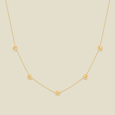 A delicate gold chain necklace featuring five small, evenly spaced textured peony flower motifs set with tiny clear stones, displayed on a solid light beige background.