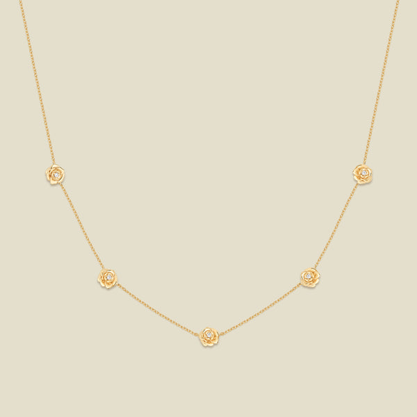 A delicate gold chain necklace featuring five small, evenly spaced textured peony flower motifs set with tiny clear stones, displayed on a solid light beige background.