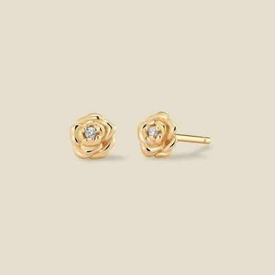 A pair of gold peony stud earrings, one shown from the front and the other from the side, each featuring a small clear gemstone at the center, set against a solid light beige background.