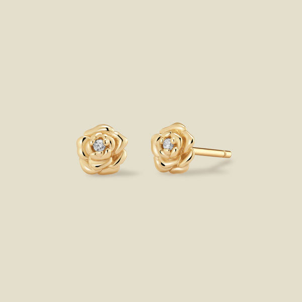 A pair of gold peony stud earrings, one shown from the front and the other from the side, each featuring a small clear gemstone at the center, set against a solid light beige background.