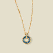 A gold chain necklace with a circular pendant featuring blue-green baguette-cut stones around a hollow center, displayed against a solid light beige background.
