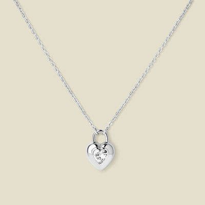 A dainty silver pendant necklace with a heart padlock charm, featuring a heart shaped clear stone in the center, set against a beige backdrop.