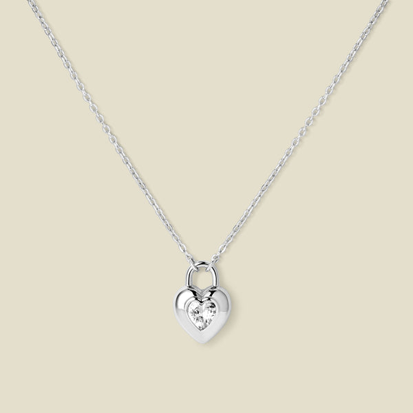 A dainty silver pendant necklace with a heart padlock charm, featuring a heart shaped clear stone in the center, set against a beige backdrop.