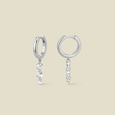 A pair of silver hoop earrings with a column of three clear round crystals hanging vertically from each, shown on a solid beige background.
