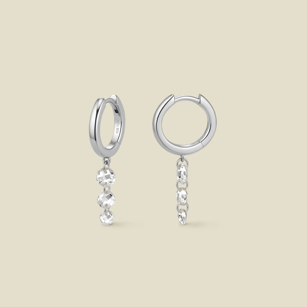A pair of silver hoop earrings with a column of three clear round crystals hanging vertically from each, shown on a solid beige background.
