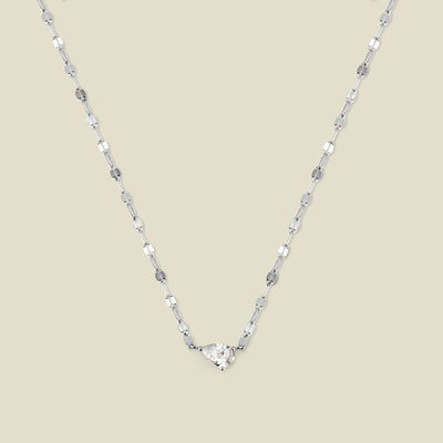 A dainty textured silver chain necklace featuring a solitaire tear-drop clear stone set sideways in the center, shown against a solid beige background.