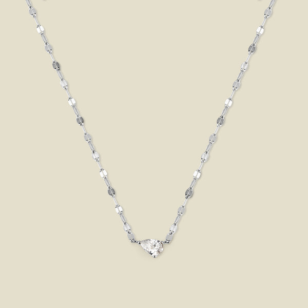 A dainty textured silver chain necklace featuring a solitaire tear-drop clear stone set sideways in the center, shown against a solid beige background.