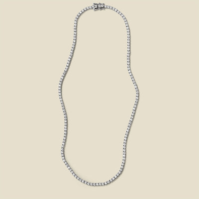 A silver prong-set tennis necklace, set against a solid beige background.