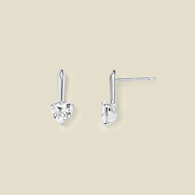 A pair of silver drop earrings with heart-shaped clear gemstones, shown from front and side angles on a solid beige background.