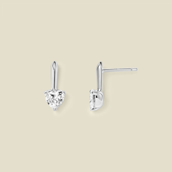 A pair of silver drop earrings with heart-shaped clear gemstones, shown from front and side angles on a solid beige background.