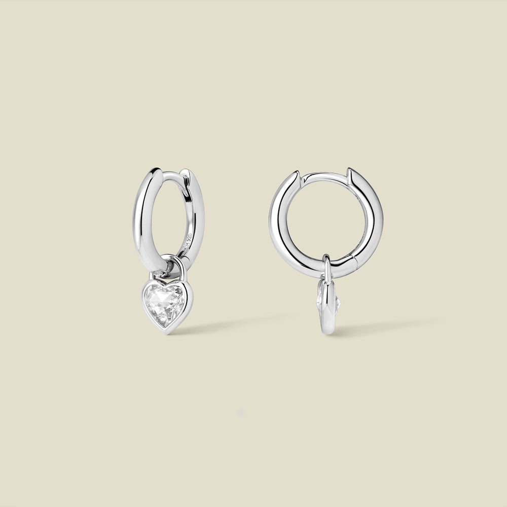 Silver hoop earrings with a dangling heart-shaped clear gemstone on a beige background