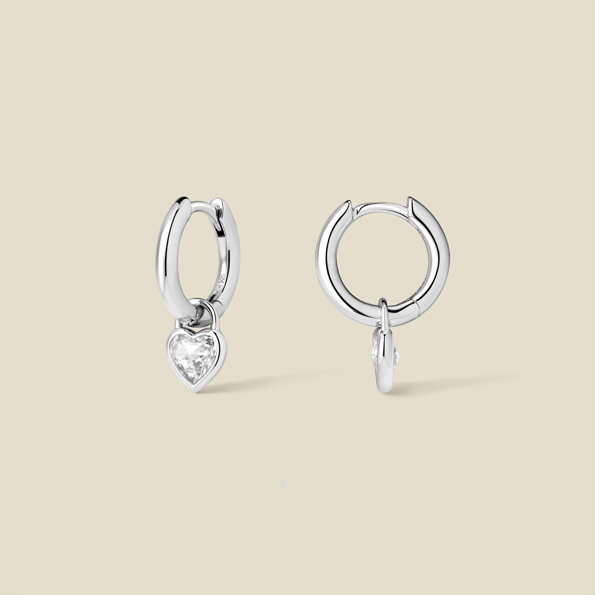 Silver hoop earrings with a dangling heart-shaped clear gemstone on a beige background