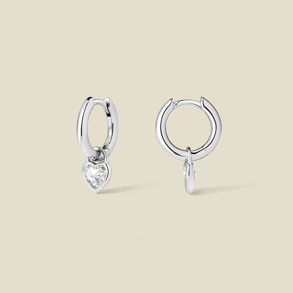 Silver hoop earrings with a dangling heart-shaped clear gemstone on a beige background