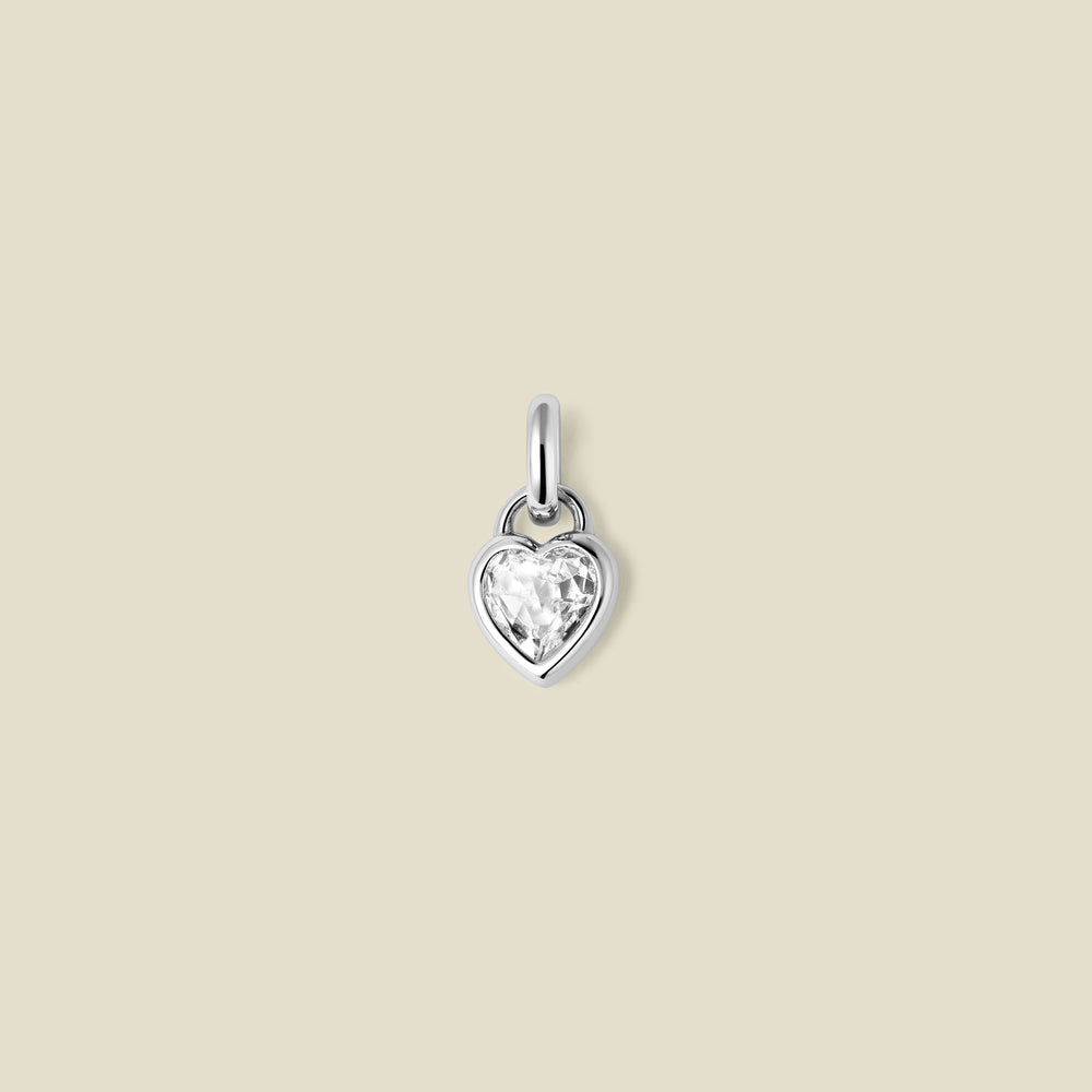 A small silver heart-shaped charm with a clear faceted heart-shaped gemstone, centered on a solid light beige background.