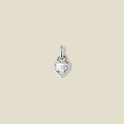 A small silver heart-shaped charm with a clear faceted heart-shaped gemstone, centered on a solid light beige background.
