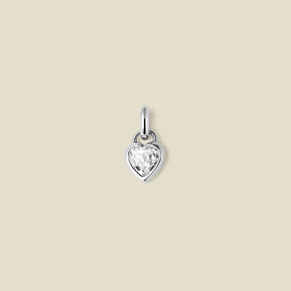 A small silver heart-shaped charm with a clear faceted heart-shaped gemstone, centered on a solid light beige background.