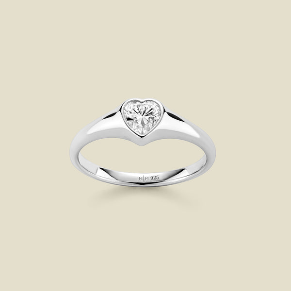 A heart-shaped clear gemstone signet ring in a silver setting, centered on a light beige background. 