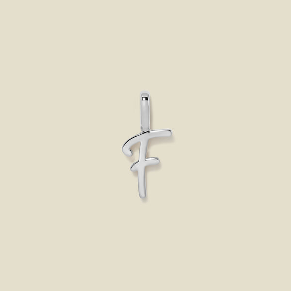 A polished silver initial pendant charm shaped like the letter 'F' in a cursive script, centered on a solid light beige background.