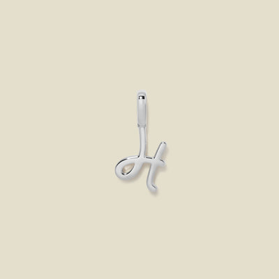 A silver pendant charm with the initial 'H' in a script font, set against a solid beige background.