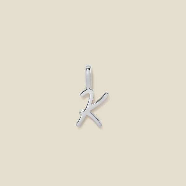 A polished silver initial pendant charm shaped like the letter 'K' in a cursive script, centered on a solid light beige background.