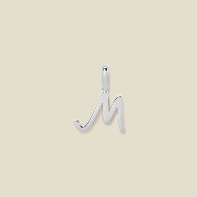 A polished silver initial pendant charm shaped like the letter 'M' in a cursive script, centered on a solid light beige background.