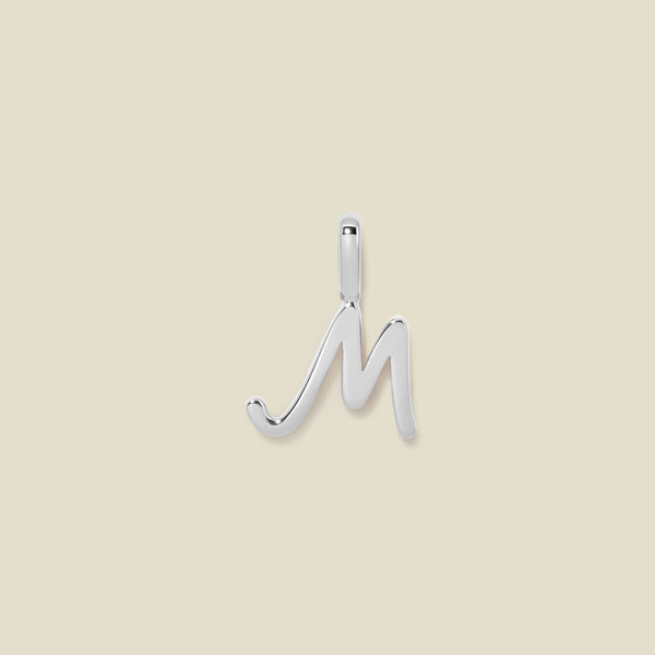 A polished silver initial pendant charm shaped like the letter 'M' in a cursive script, centered on a solid light beige background.