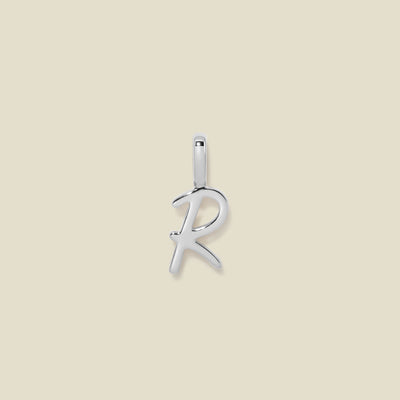 A polished silver initial pendant charm shaped like the letter 'R' in a cursive script, centered on a solid light beige background.