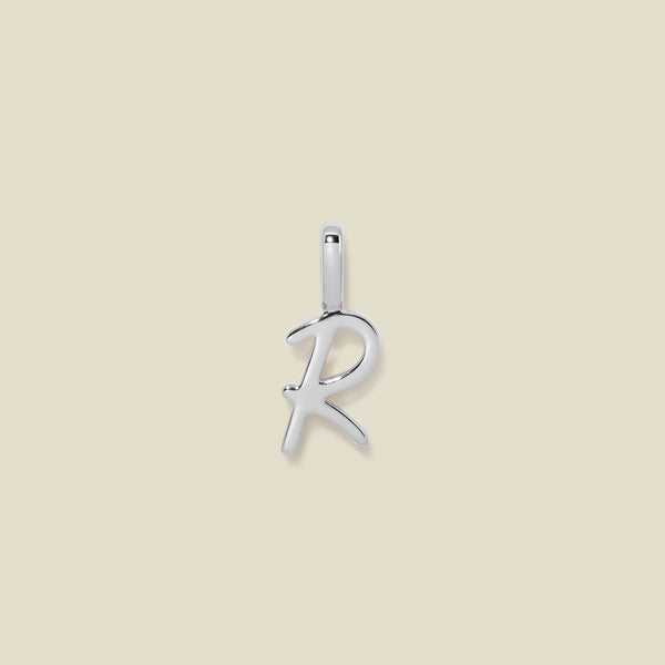 A polished silver initial pendant charm shaped like the letter 'R' in a cursive script, centered on a solid light beige background.