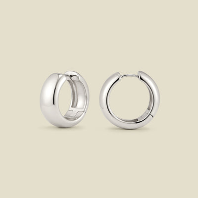 A pair of wide, polished silver hoop earrings standing up right at two different angles, set against a solid beige background.