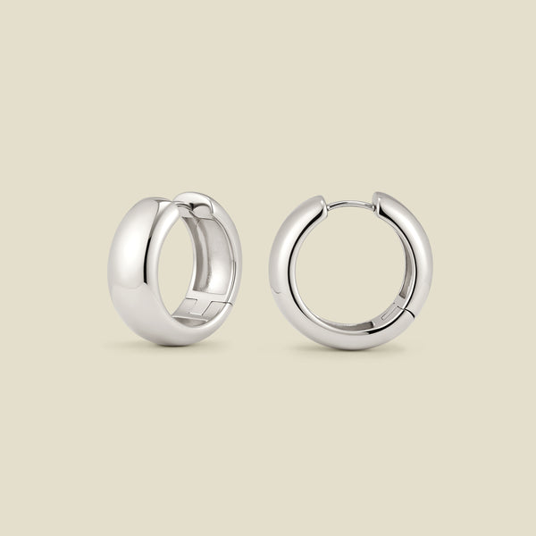 A pair of wide, polished silver hoop earrings standing up right at two different angles, set against a solid beige background.