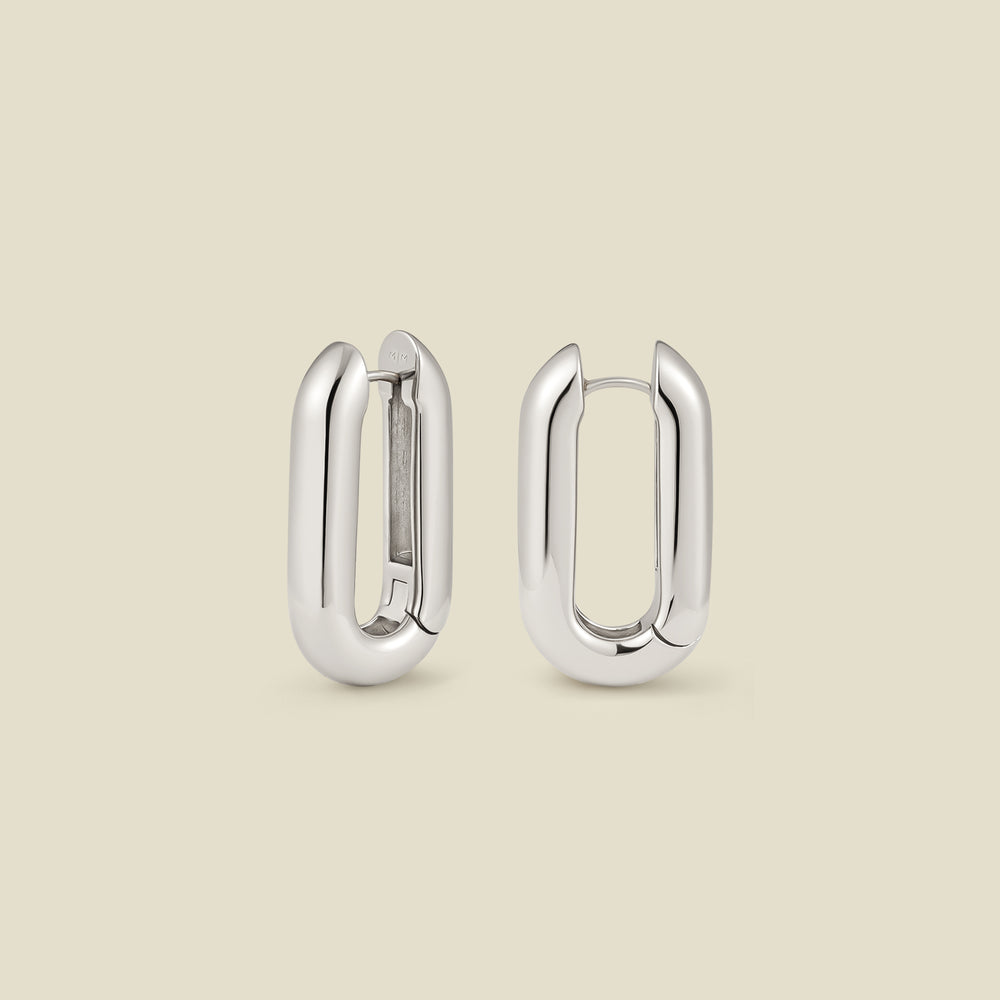 A pair of wide, polished silver paperclip shaped earrings standing up right and shown at two different angles, set against a solid beige background.