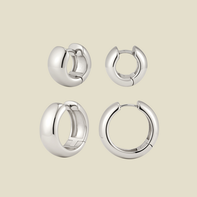 Two sets of polished silver huggie hoop earrings in two sizes, shown from different angles on a light beige background.