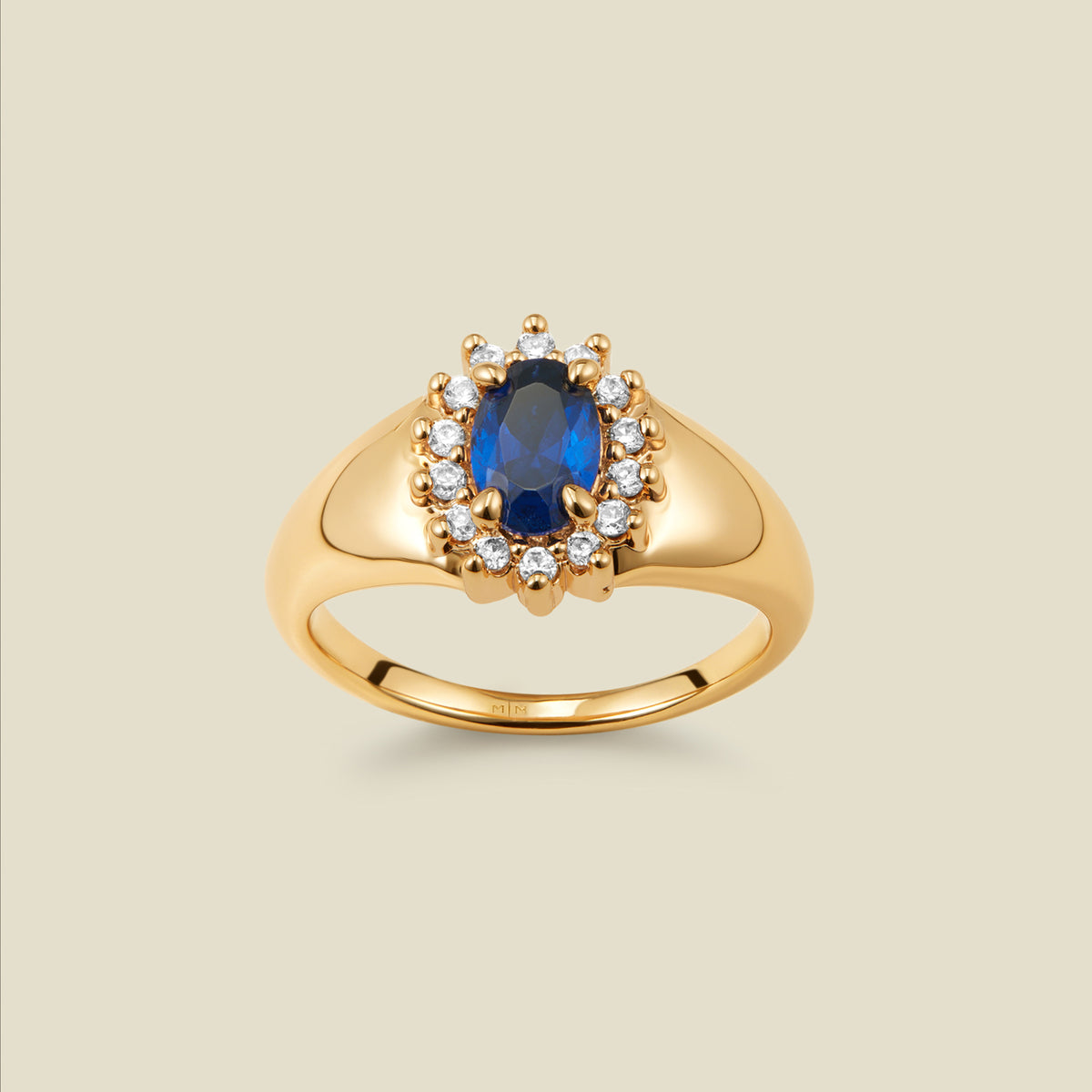 A gold signet ring with an oval blue stone framed by a halo of small clear CZ stones, on a beige background.