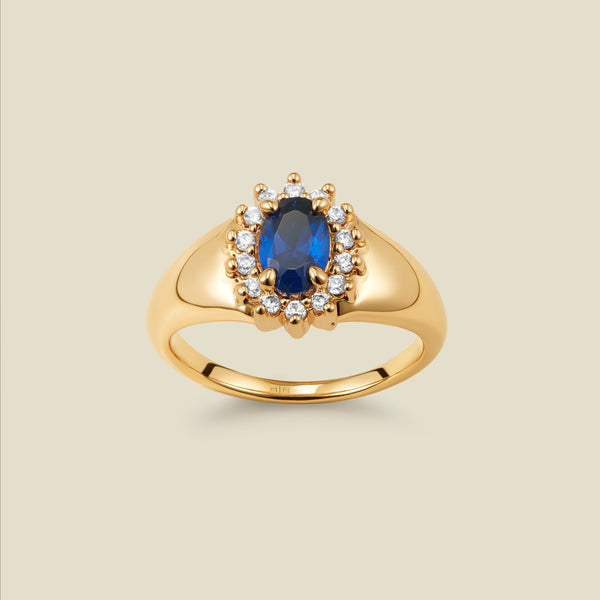 A gold signet ring with an oval blue stone framed by a halo of small clear CZ stones, on a beige background.