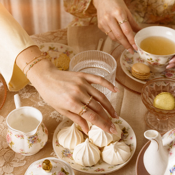 Close-up of two woman's hands reaching for food at a tea set, wearing gold rings and bracelets.