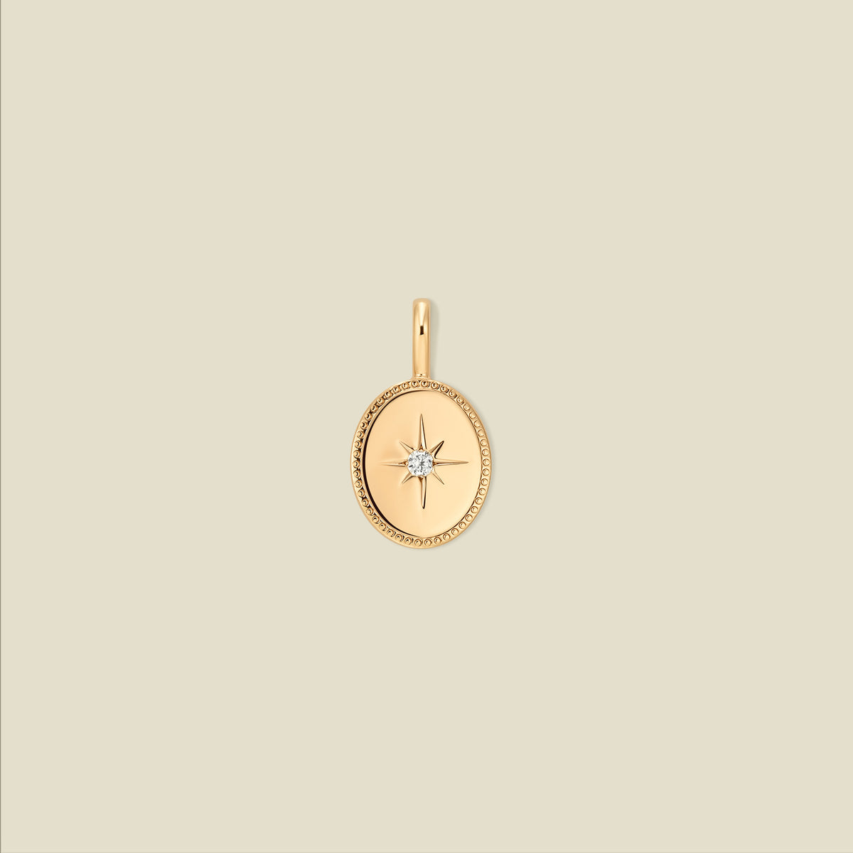 A gold oval pendant featuring a bezel-set starburst motif with a central clear gemstone, shown against a solid light beige background.