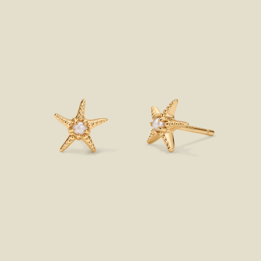 A pair of gold, textured starfish stud earrings with a pearl in the center of each, set against a solid beige background.