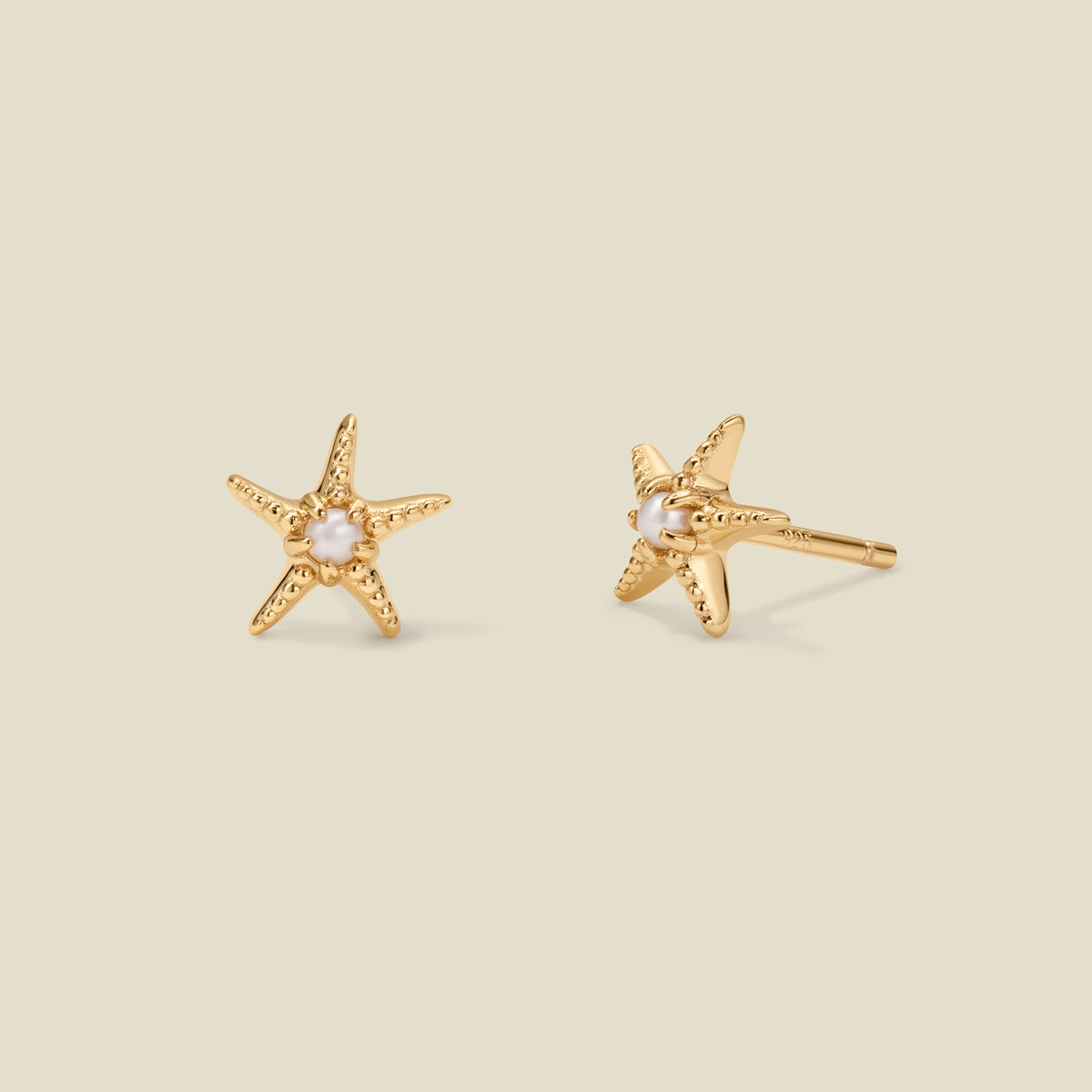 A pair of gold, textured starfish stud earrings with a pearl in the center of each, set against a solid beige background.
