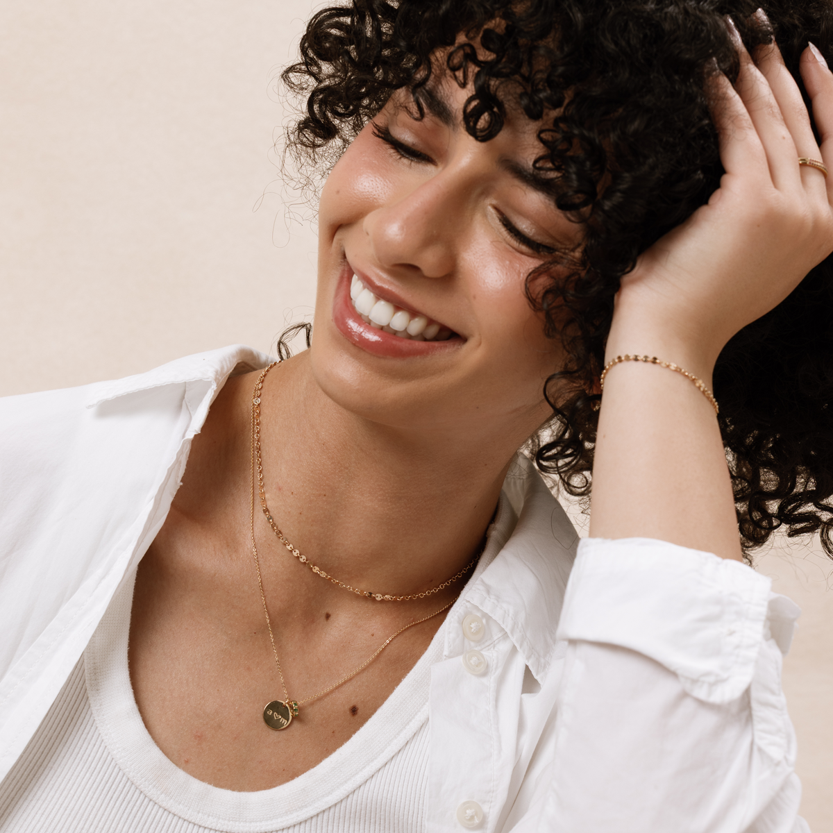 A woman wearing two layered gold necklaces, one with a round, flat pendant, and a gold chain bracelet, paired with a white linen top over a tank top and set against a beige background.