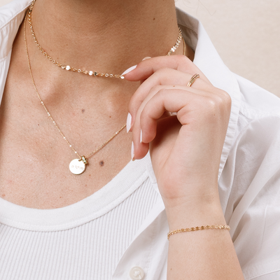 A close-up of a person wearing two layered gold necklaces, one with a round, flat pendant and small ring charm, with gold rings and a gold bracelet.