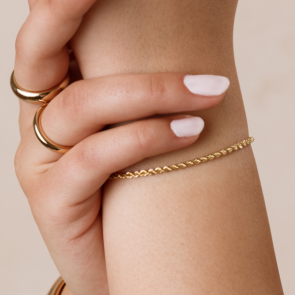 A close-up of a person's wrist wearing a rope textured chain bracelet, with two polished gold rings against a solid beige background.